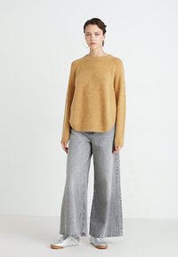 Mustard-colored knit sweater with a round neckline, paired with wide-leg gray jeans and silver sneakers. Model stands against a plain background.