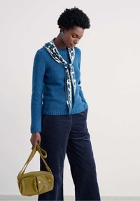 Egy nő kék pulóvert és sálat visel, olíva zöld táskát tart, egyik kezét zsebébe dugva áll egy sima háttér előtt.