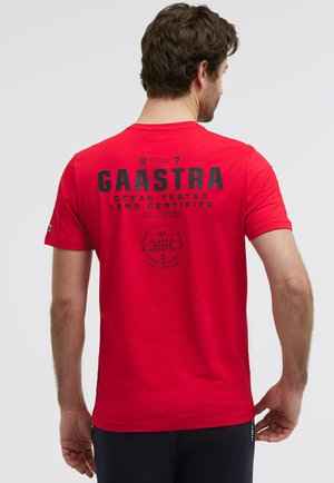Mann trägt rotes Gaastra-T-Shirt mit schwarzem, nautisch inspiriertem Text und Logos auf der Rückseite, steht mit dem Rücken zur Kamera vor einem hellen, einfarbigen Hintergrund.