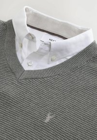 Suéter gris de punto con escote en V, que cuenta con una camisa blanca con cuello debajo. Logo bordado en el pecho. Acabado de tela texturizada.