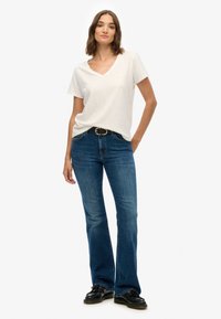 Camiseta blanca de cuello en V, vaqueros acampanados de mezclilla, cinturón negro con hebilla redonda, zapatos negros, modelo posando de manera casual contra un fondo sencillo.