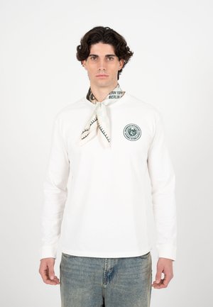 Młody mężczyzna o ciemnych, kręconych włosach, ubrany w białą koszulę z długim rękawem z okrągłym logo oraz jasny szalik zawiązany wokół szyi.