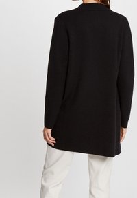 Lång svart cardigan i mjukt tyg, med krage och långa ärmar, rak skärning och minimalt synliga sömmar.