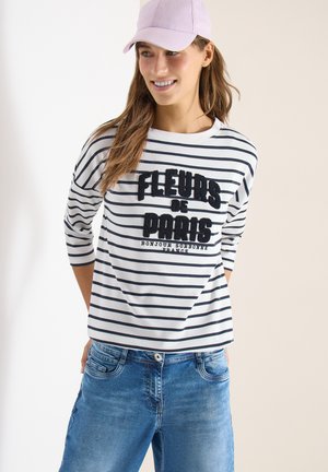 Mujer sonriente, vistiendo una camiseta a rayas con la inscripción "Fleurs de Paris", jeans azules y una gorra de béisbol rosa claro, de pie con las manos detrás de la espalda.