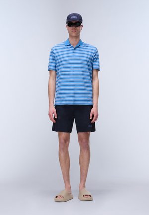 Mann stehend, trägt blau gestreiftes Poloshirt, schwarze Shorts, beige Pantoffelsandalen, Navy-Kappe und schwarze Sonnenbrille, vor einem schlichten hellen Hintergrund.