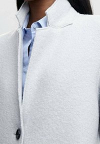 Lichtgrijze wollen blend jas met een hoge kraag, voorzien van een enkele zwarte knoopsluiting en een textuurafwerking, gedragen over een blauw shirt.