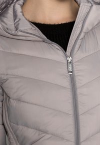 Svetle šedá prošívaná bunda zapnutá přes černý pletený top, který odhaluje dolní část krku a horní část hrudníku.