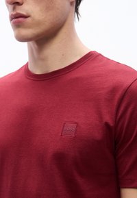 Νεαρός άνδρας που φοράει απλό κόκκινο T-shirt με στρογγυλή λαιμόκοψη και μικρό κεντητό λογότ υπο "BOSS" στο αριστερό στήθος.