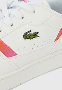 Λευκό αθλητικό παπούτσι με λεία επιφάνεια, που διαθέτει ροζ και πορτοκαλί λεπτομέρειες, διάτρητο σχέδιο και πράσινο κεντημένο λογότυπο κροκόδειλου.