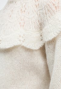 Gros plan sur un tissu tricoté doux de couleur crème avec un motif délicat en dentelle et un détail de bord ondulé.