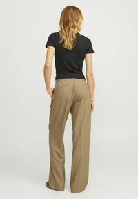 Un t-shirt noir ajusté et un pantalon beige à jambe large sont portés. Le pantalon présente deux poches arrière et une finition lisse et ajustée.