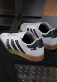White sneakers with black stripes, textured fabric, and a rubber sole. The heel features a black accent and a "CORDURA" label.