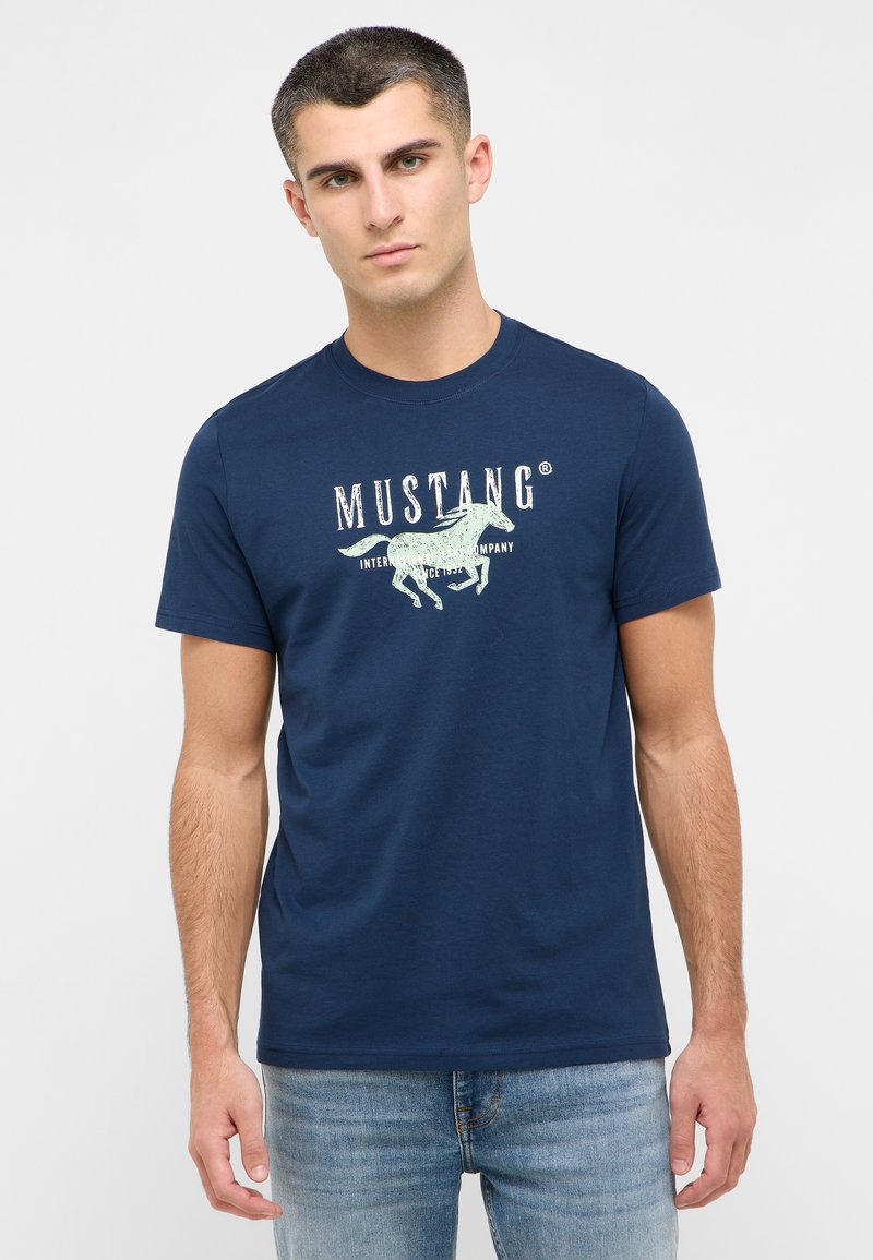 T-shirt en coton bleu marine avec un col rond. Présente un graphique de cheval et le mot "MUSTANG" en grandes lettres blanches sur le devant.