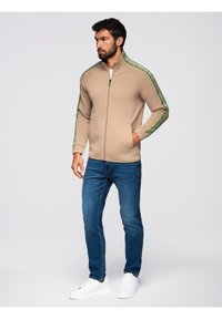 Giacca beige con zip e strisce laterali verdi, dotata di colletto alto e tasche, indossata con jeans blu e sneaker bianche.