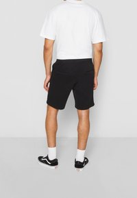 Homme portant un short noir, un t-shirt blanc, des chaussettes blanches et des baskets noires, se tenant devant un mur gris clair uni.