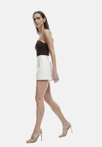 Top marrón sin mangas, mini falda blanca texturizada y sandalias doradas de tacón alto con tiras. Modelo caminando con el cabello largo suelto.