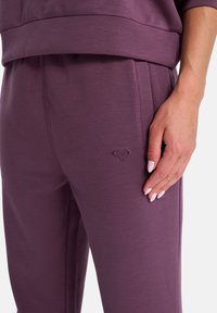 Lila joggingbyxor gjorda av mjukt tyg, med en subtil präglad logotyp, elastisk midja och sidofickor. Slät textur med en avslappnad passform.