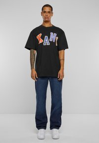 Black oversized t-shirt with colorful "XANI" text in orange, purple, and blue. Paired with blue denim jeans and white sneakers.