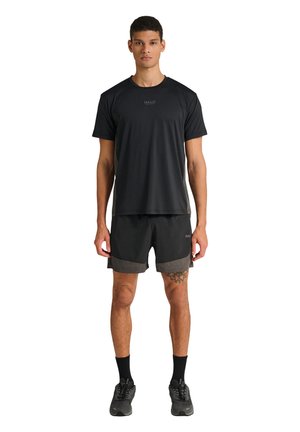 Młody mężczyzna stojący przodem, ubrany w czarną odzież sportową z logo "HALO", czarne skarpetki i czarne buty do biegania, na jednolitym białym tle.