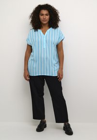 Tunique bleu clair à manches courtes avec des rayures blanches verticales et un col en V ; associée à un pantalon noir à jambes larges et des chaussures noires.