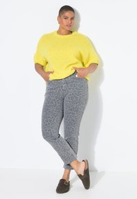 Suéter de punto amarillo con mangas cortas, jeans grises con estampado de leopardo y zapatos marrones sin cordones con detalle de hebilla, con las manos en los bolsillos.