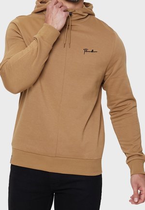 Homme ajustant le cordon de serrage d'un sweat à capuche beige avec un logo brodé noir, portant un pantalon noir sur un fond clair uni.