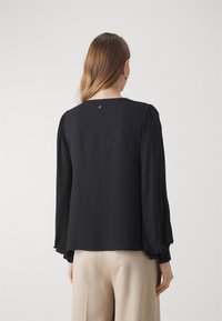 Black long-sleeve blouse with gathered shoulders and cuffs, featuring a round neckline and a back button detail, paired with light beige trousers.