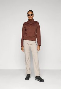 Sieviete, kas valkā brūnu ceturtdaļas rāvējslēdzēja džemperi, bēšas bikses, melnas pārgājienu zābakus un saulesbrilles, stāv pret vienkāršu gaiši pelēku fonu.