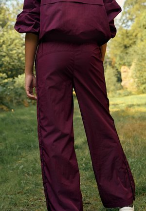 Bordo värvi treeningpüksid, millel on sile tekstuur, laiad sääred, külgsed tõmblukud ja kontrastsed mustad aktsendid, näidatud tagant rohelise tausta ees.