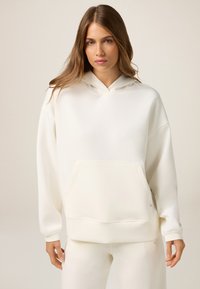 Sudadera blanca con capucha, con un bolsillo frontal, corte relajado y puños acanalados. Textura suave y diseño minimalista con un sutil detalle de marca.