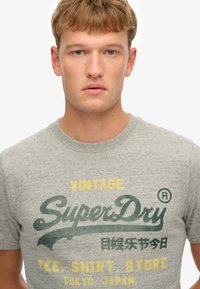 Camiseta gris de manga corta con el logo "SuperDry" en estampado verde y amarillo, cuello redondo y tela suave y texturizada.