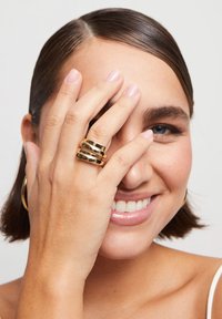 Conjunto de anillos de oro que presenta tres bandas apiladas con un acabado liso y brillante. La mano está suavemente situada cerca de la cara, parcialmente oscureciéndola.