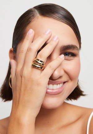 Conjunto de anillos de oro que presenta tres bandas apiladas con un acabado liso y brillante. La mano está suavemente situada cerca de la cara, parcialmente oscureciéndola.