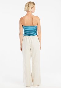 Vrouw met kort blond haar kijkt weg, gekleed in een blauw-wit gestreept strapless top en losse beige broek tegen een witte achtergrond.