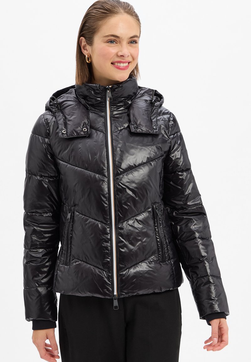 Femme souriante portant une veste matelassée noire brillante avec un col montant et des poches zippées, se tenant contre un fond blanc.