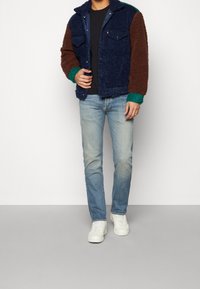Chupa de forro polar con bloques de color azul marino, marrón y verde, que presenta un cierre de botones y dos bolsillos en el pecho, combinado con jeans de un azul claro y zapatillas blancas.