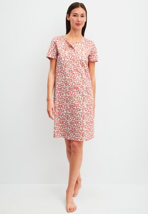 Vrouw staat op blote voeten, draagt een knielange jurk met korte mouwen en een roze-wit bloemmotief, tegen een effen lichte achtergrond.