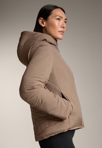 Brun vadderad jacka med huva, med sidofickor och en mjuk, texturerad fåll. Lätt tyg med synliga sömnadsdetaljer.