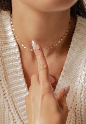 Dedo con uñas cuidadas tocando un delicado collar de cadena dorada en una persona que lleva un suéter de punto color crema.