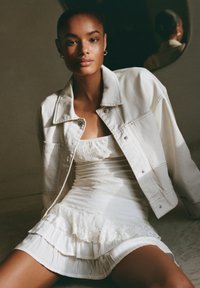Veste en denim blanche sur une mini robe blanche texturée avec un ourlet froncé. Tissu doux, corsage ajusté, et détails subtils en dentelle visibles.