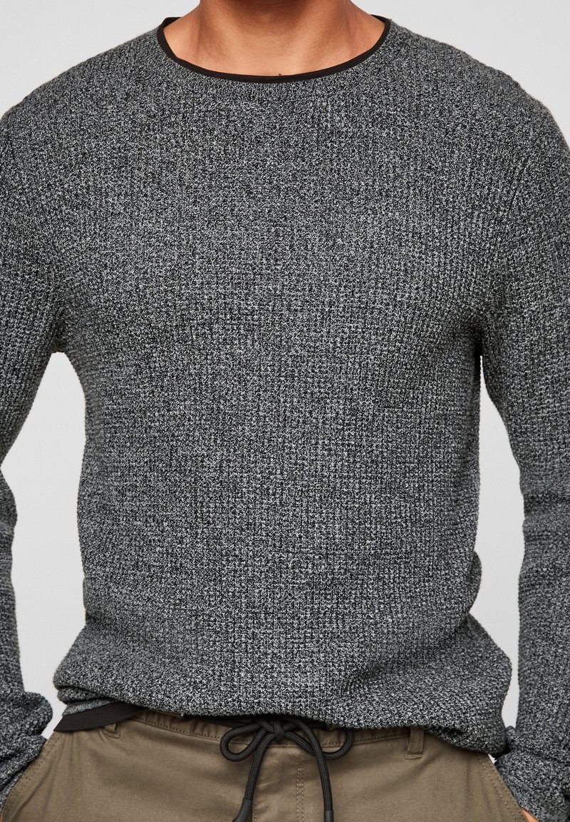 Pull gris texturé avec un col rond et un bord noir contrastant ; le tissu présente un motif tricoté et des poignets ajustés.