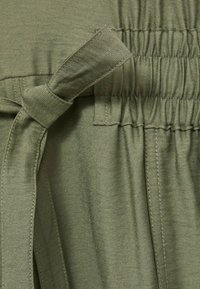 Groene stof met een gestructureerde afwerking. Heeft een gestrikte taille met een knoop en een geplooide elastische afwerking aan de bovenkant voor een getailleerd effect.