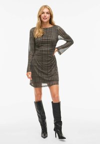 Robe évasée à motif écossais brun avec de longues manches transparentes. Col haut et longueur au-dessus du genou. Associée à des bottes noires ajustées montantes.