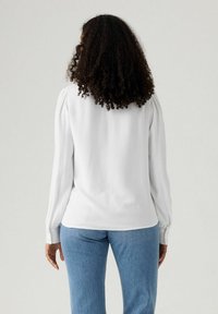 Blusa bianca a maniche lunghe con spalle a palloncino e polsini abbottonati, abbinata a jeans in denim blu. Tessuto liscio, design semplice senza motivi.
