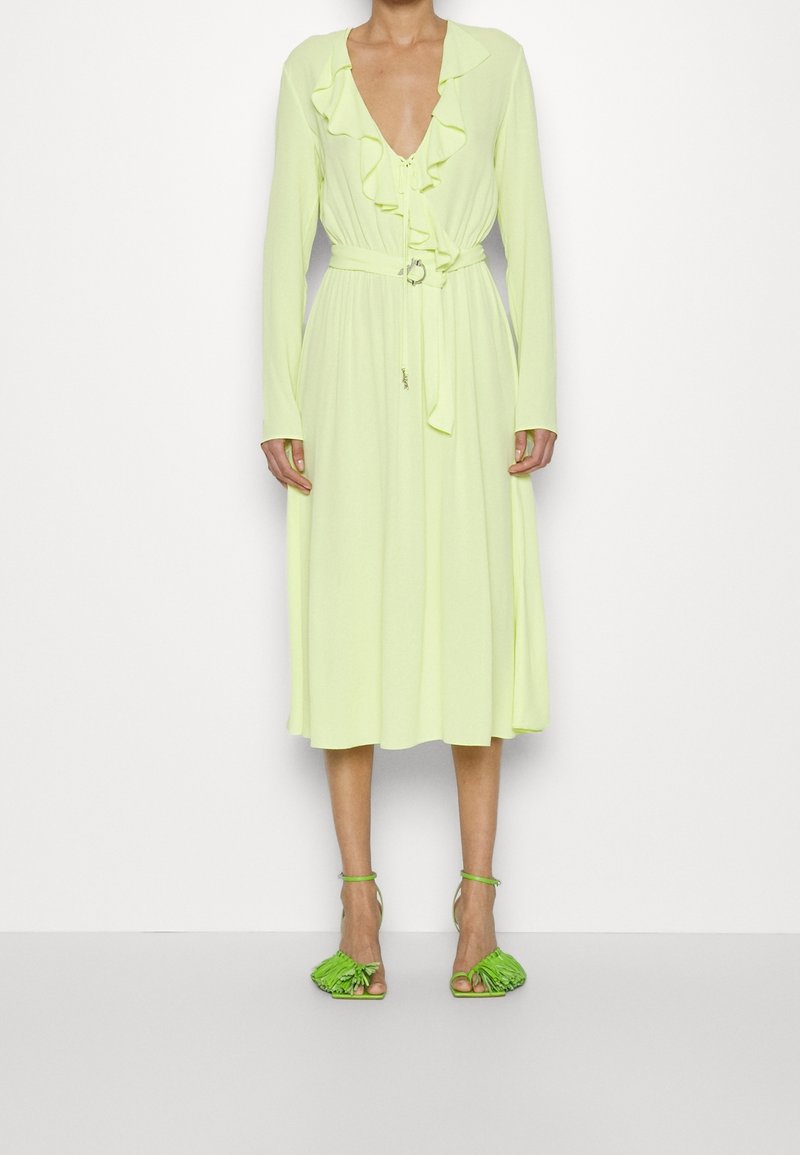 Femme portant une robe midi jaune clair à manches longues avec volants et ceinture, associée à des talons ouverts à franges vert vif.
