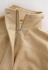 Col de veste beige avec fermeture éclair argentée partiellement ouverte et trois boutons-pression sur tissu doux.