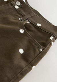 Bruine spijkerbroek met witte stippen, voorzien van een voorzak, zilveren klinknagels en een knoopsluiting op een lichte achtergrond.