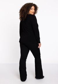 Schwarzes langärmliges Oberteil mit gerafften Ärmeln, kombiniert mit schwarzen Schlaghosen. Glatte Textur, figurbetonte Silhouette und minimalistisches Design.