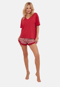 Czerwona bluzka z dekoltem w serek wykonana z miękkiego materiału, zestawiona z wzorzystymi szortami w białe serca na czerwonym tle. Luźny, swobodny fason.
