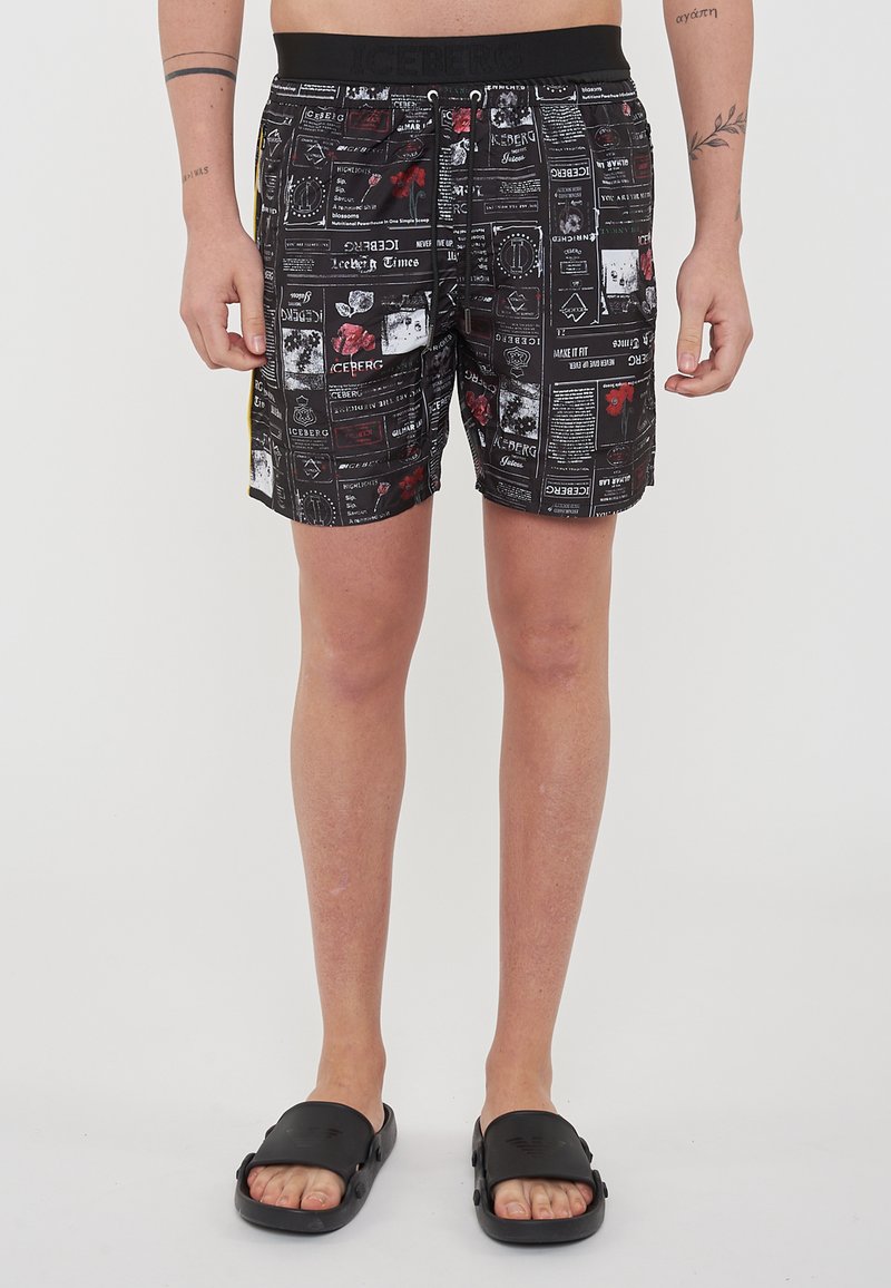 Pantaloni da bagno neri con stampe grafiche colorate, vita elastica con cordino e tasche laterali. Indossati con sandali neri a ciabatta.